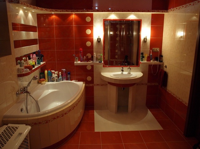 Wykończona łazienka z narożną wanną, umywalką na postumencie i czerwono-białymi płytkami na ścianach i podłodze, oświetlona punktowymi lampami.