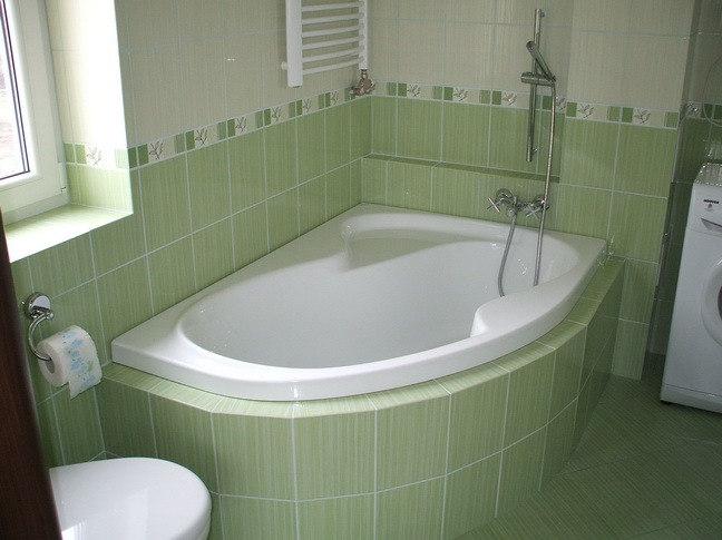 Narożna wanna obudowana zielonymi płytkami z dekoracyjnym paskiem, bateria wannowa z słuchawką prysznicową, grzejnik drabinkowy i pralka w tle.