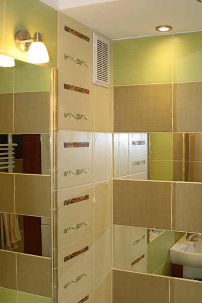 Wykończona łazienka z kremowo-brązowymi płytkami z motywem bambusa, wentylator w rogu i odbicie umywalki w lustrzanej płytce.