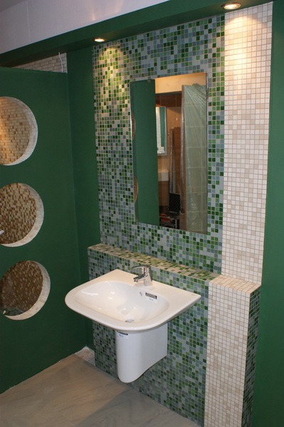 Łazienka z umywalką podwieszaną, obudowana zieloną mozaiką z kwadratowych płytek, lustro na ścianie z mozaiką, zielona ściana z okrągłymi wnękami wykończonymi mozaiką.