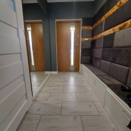 Pruzo Marek Strzała - Nowoczesny przedpokój z panelami ściennymi w odcieniach szarości, drewnianymi akcentami i białymi szafkami w zabudowie, wykończony podłogą z jasnych płytek imitujących drewno.