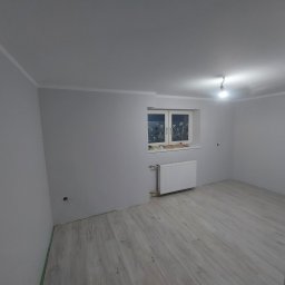 Pruzo Marek Strzała - Pokój z jasnymi panelami podłogowymi ułożonymi wzdłuż, białe ściany z listwami, okno z firankami i grzejnik pod nim, surowa żarówka na suficie.