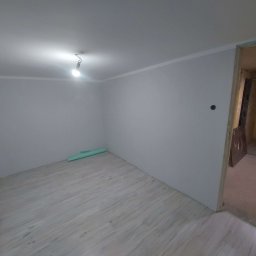 Pruzo Marek Strzała - Pokój po remoncie z jasnymi panelami podłogowymi ułożonymi wzdłuż dłuższego boku, świeżo pomalowanymi szarymi ścianami, grzejnikiem pod oknem i odkrytym otworem drzwiowym do kolejnego pomieszczenia...