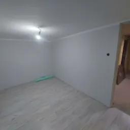 Pokój po remoncie z jasnymi panelami podłogowymi ułożonymi wzdłuż dłuższego boku, świeżo pomalowanymi szarymi ścianami, grzejnikiem pod oknem i odkrytym otworem drzwiowym do kolejnego pomieszczenia...