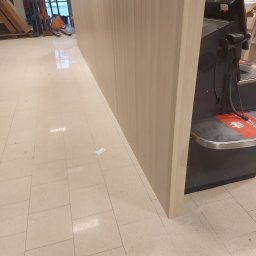 Pruzo Marek Strzała - Jasny panel z płyty meblowej zamontowany jako element lady sklepowej, widoczna podłoga z płytek w odcieniach beżu oraz fragment kasy fiskalnej z czytnikiem kodów kreskowych.