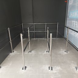 Pruzo Marek Strzała - Metalowe barierki z czarnymi pasami wyznaczające przestrzeń, ustawione na szarej podłodze w płytkach, przy czarnej ścianie i szybie z napisem.