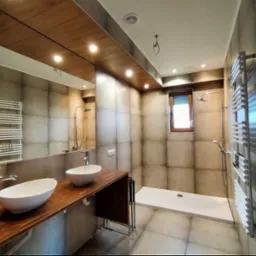 Nowoczesna łazienka z dwoma owalnymi umywalkami nablatowymi na drewnianym blacie, kabiną prysznicową typu walk-in i grzejnikiem drabinkowym. Sufit wykończony drewnianymi panelami.