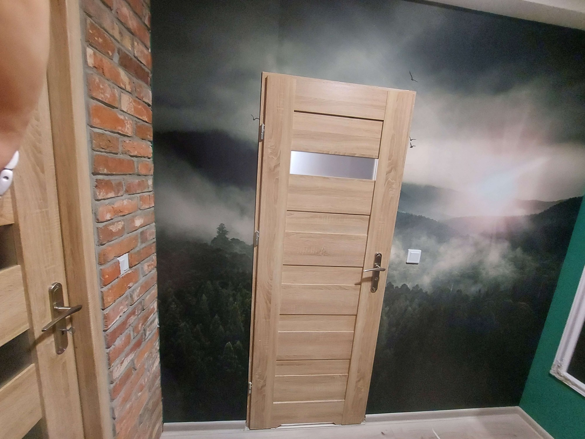 Zamontowane drzwi wewnętrzne z poziomymi panelami i szybą, widoczne na tle ściany z fototapetą leśną, sąsiadują z fragmentem ściany z cegły.