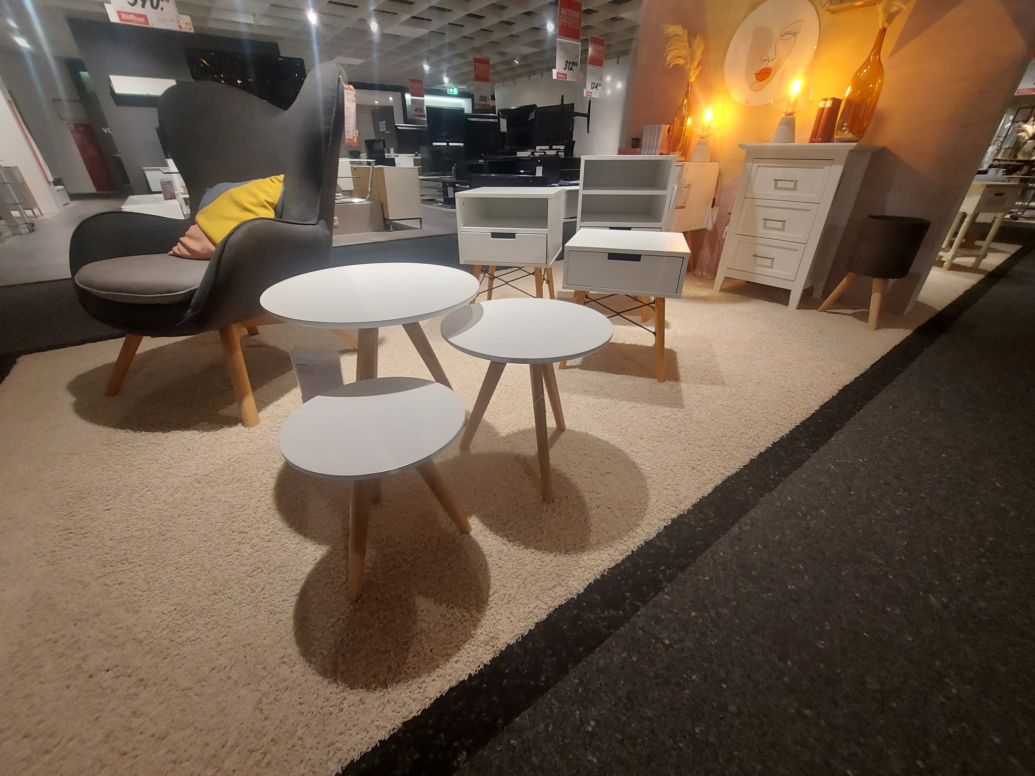 Ekspozycja mebli w salonie: zestaw trzech okrągłych stolików kawowych z białymi blatami i drewnianymi nogami, szary fotel z poduszką, biała komoda z trzema szufladami oraz szafki nocne...