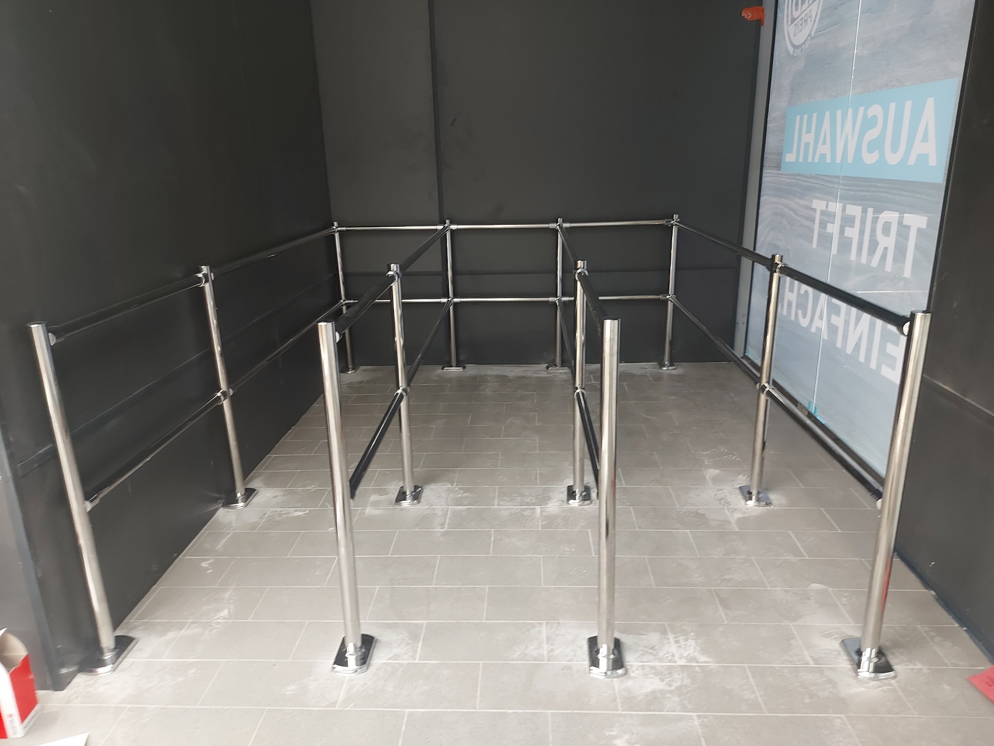 Metalowe barierki z czarnymi pasami wyznaczające przestrzeń, ustawione na szarej podłodze w płytkach, przy czarnej ścianie i szybie z napisem.