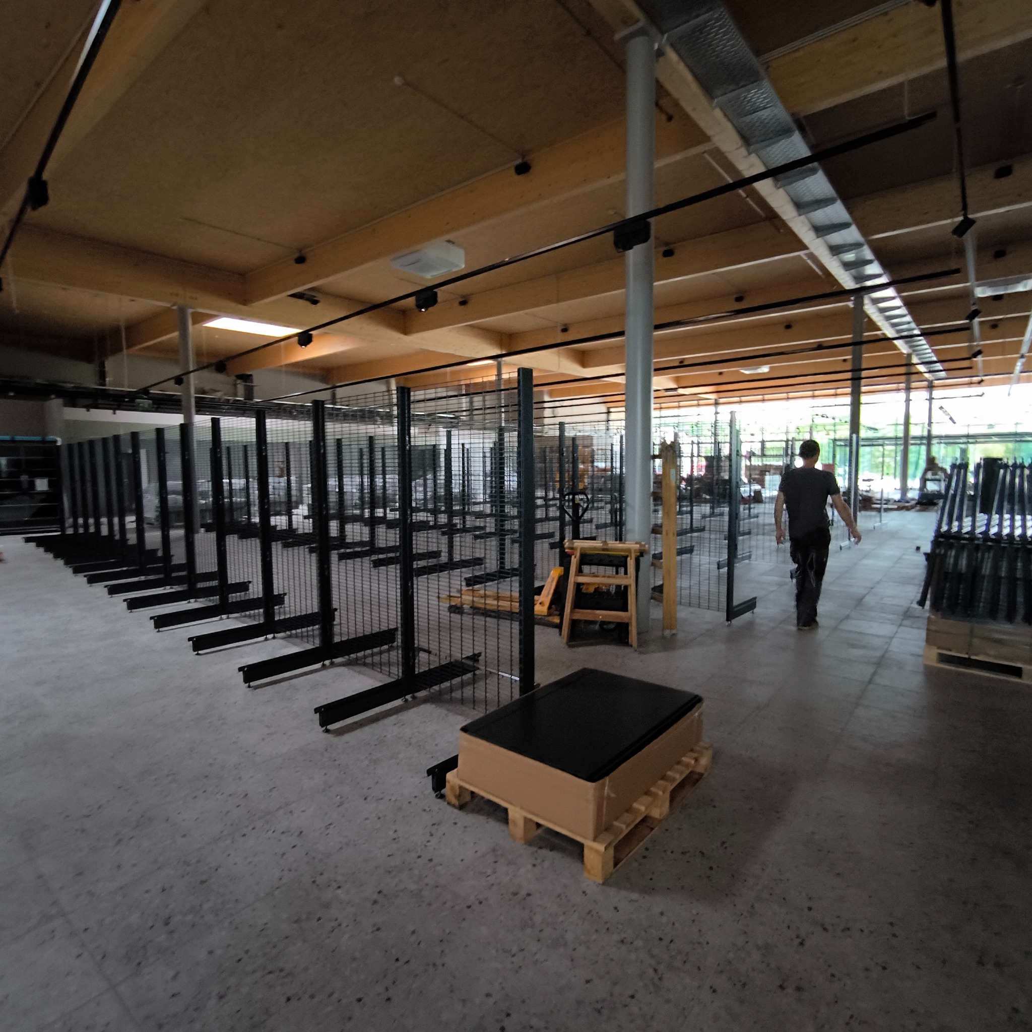 Rozległe wnętrze sklepu w trakcie wyposażania: rząd metalowych regałów z przymocowanymi półkami, drewniany sufit z widocznymi instalacjami, pracownik w oddali.