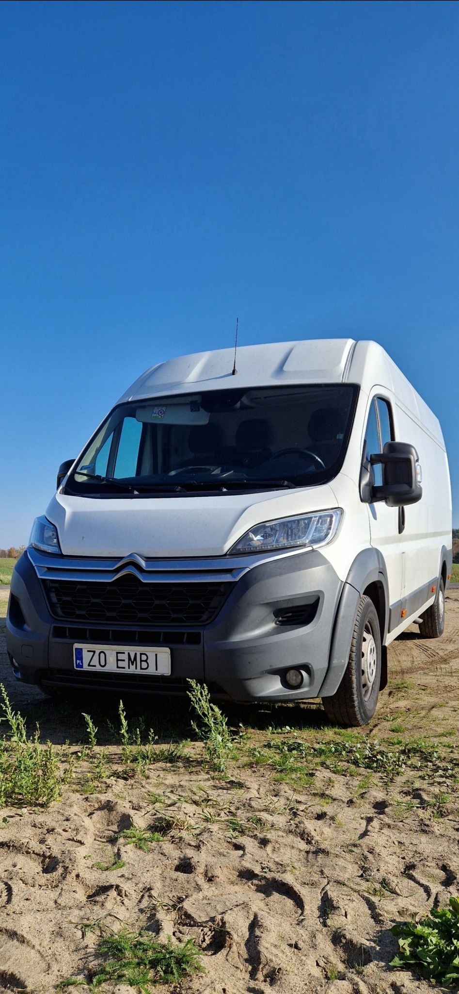 Biały samochód dostawczy marki Citroen, zaparkowany na piaszczystym podłożu z widocznymi śladami opon, z błękitnym niebem w tle.