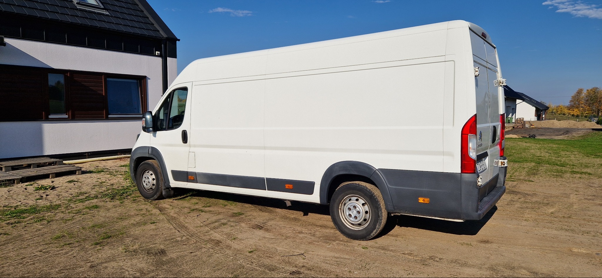 Biały furgon dostawczy marki Peugeot, zaparkowany na placu budowy z widocznymi śladami błota na oponach i karoserii, z domem w tle.