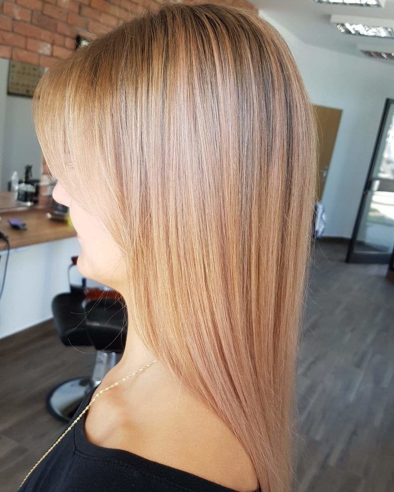 Profil kobiety z długimi, prostymi włosami w odcieniu truskawkowego blondu z delikatną grzywką, widoczna złota biżuteria na szyi, w tle ceglana ściana i fotel fryzjerski.