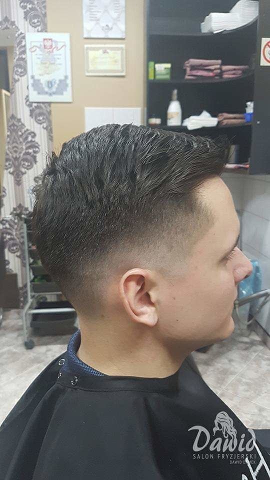 Męska fryzura z cieniowaniem, widok z profilu, klient ubrany w pelerynę fryzjerską, w tle półki z kosmetykami i dyplomy na ścianie.