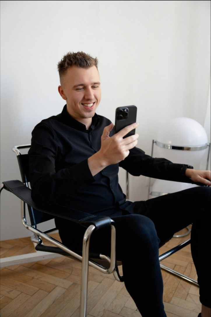 Uśmiechnięty młody mężczyzna w czarnej koszuli siedzi na skórzanym krześle Marcel Breuer, przeglądając coś na smartfonie z potrójnym aparatem.