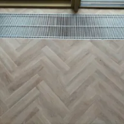 Jasne panele ułożone w jodełkę, fragment grzejnika pod oknem, zielony znacznik na podłodze.