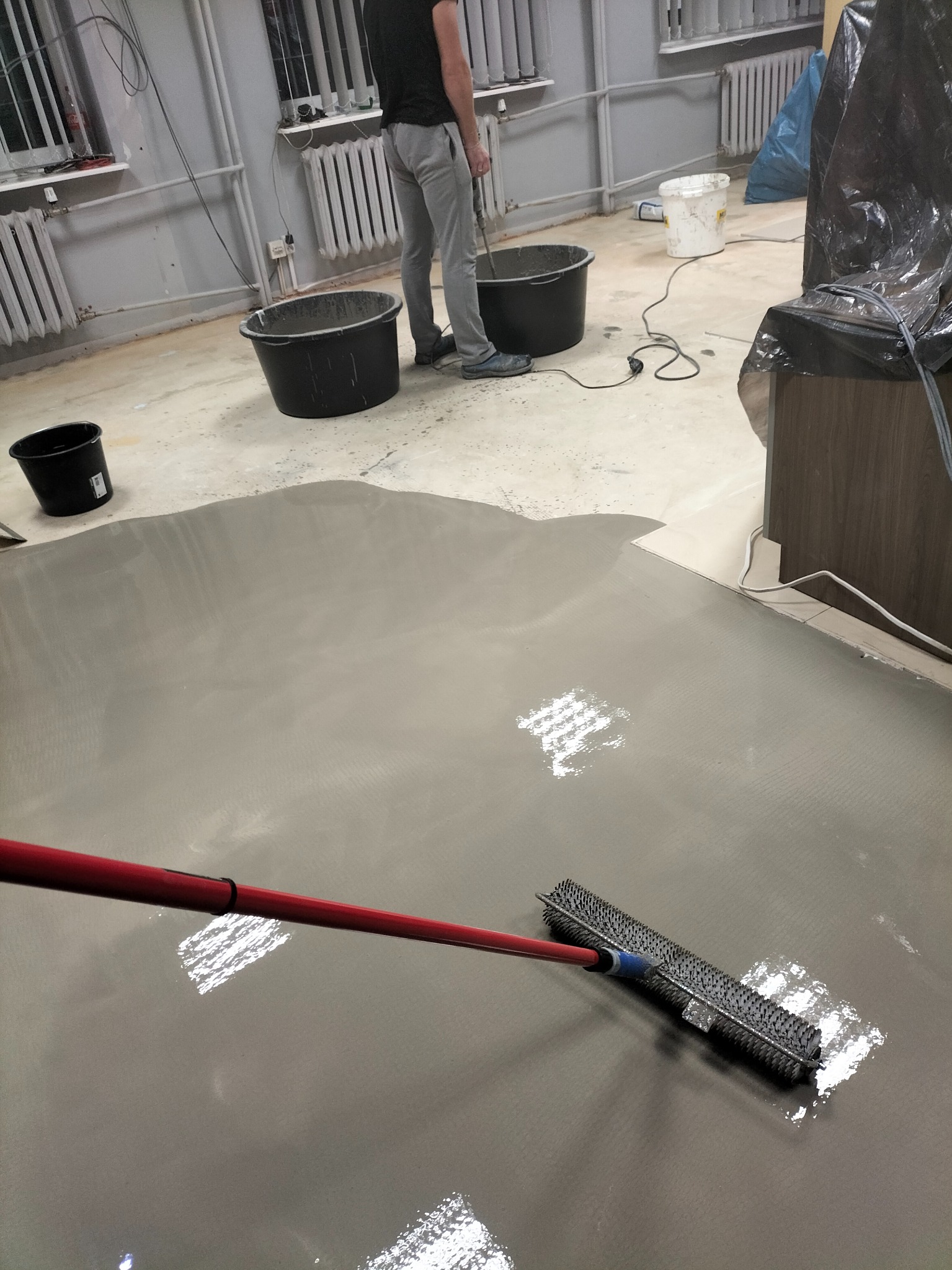 Wylewka samopoziomująca rozprowadzana wałkiem kolczastym w pomieszczeniu z grzejnikami i wiadrami z zaprawą.