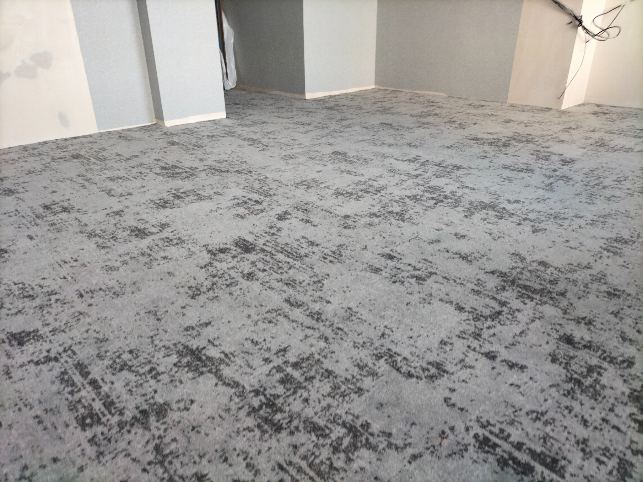 Szara wykładzina dywanowa o abstrakcyjnym wzorze w nowym pomieszczeniu z pomalowanymi ścianami i wystającymi kablami.