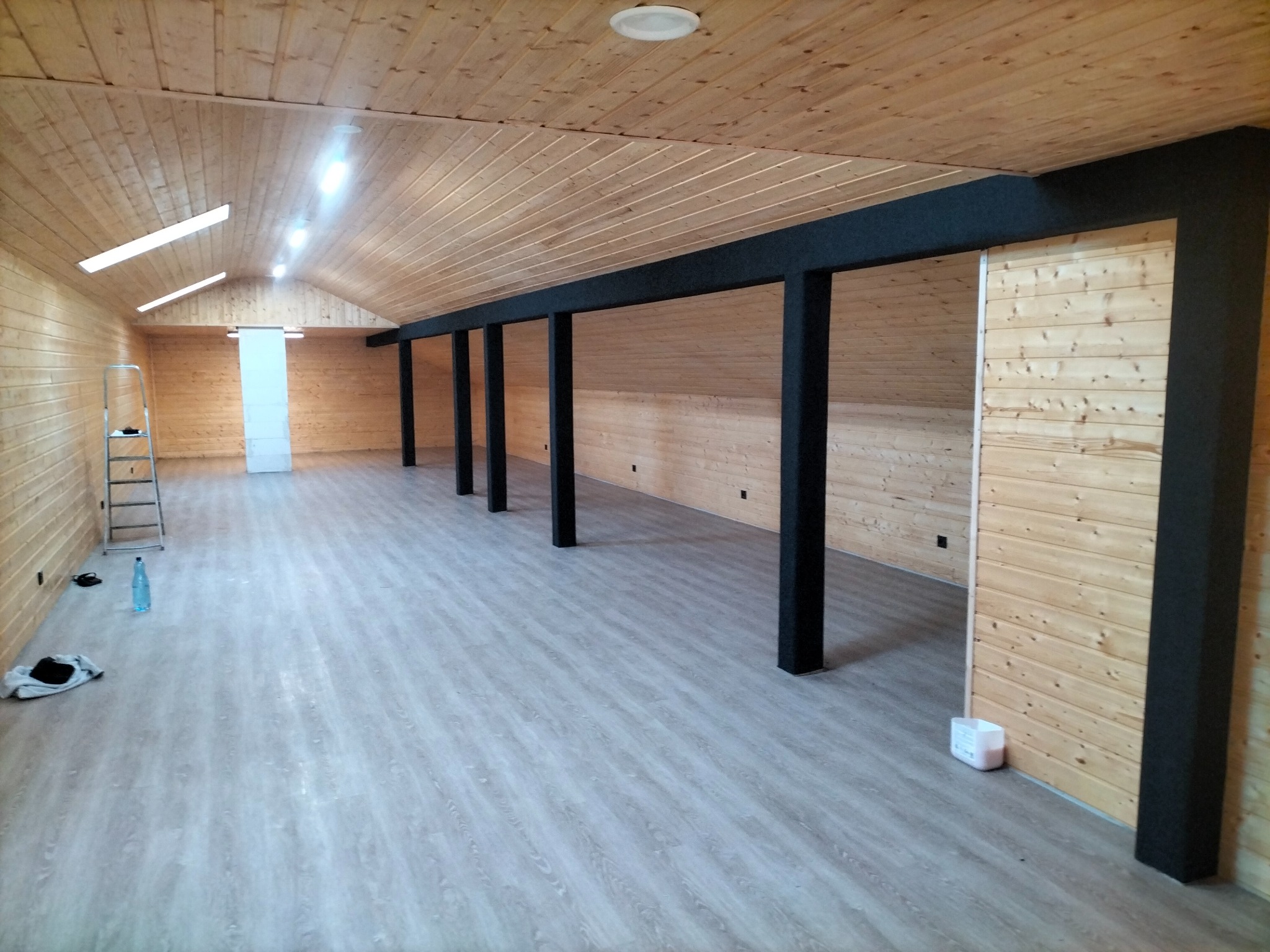 Wykończone poddasze z panelami podłogowymi w odcieniach szarości, drewnianą boazerią na ścianach i suficie, czarne belki konstrukcyjne, drabina oparta o ścianę w oddali, butelka wody i biały...