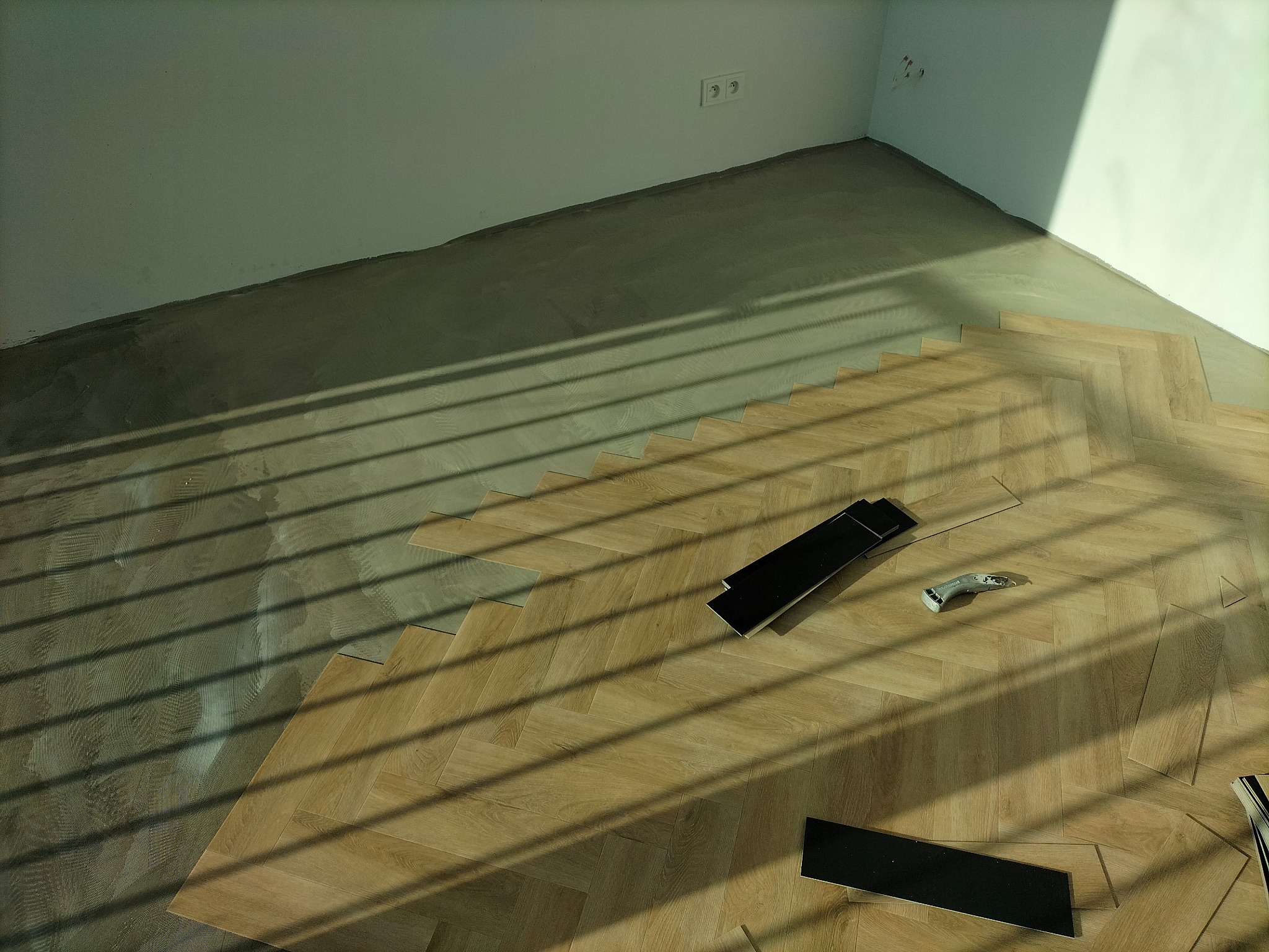 Fragment podłogi w jodełkę podczas układania paneli, widoczne przycięte panele, narzędzie do cięcia i cień rzucany przez żaluzje.