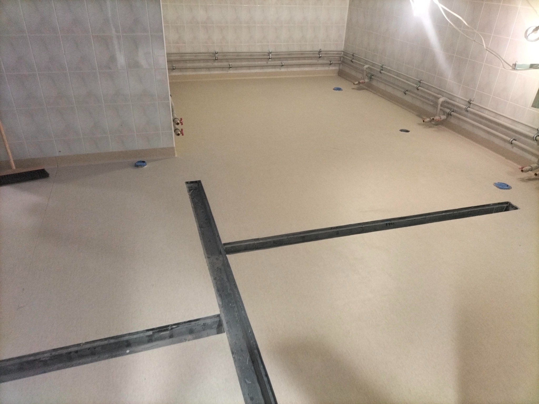 Przygotowane podłoże pod wykładzinę w pomieszczeniu z instalacją sanitarną na ścianach i metalową konstrukcją w podłodze.