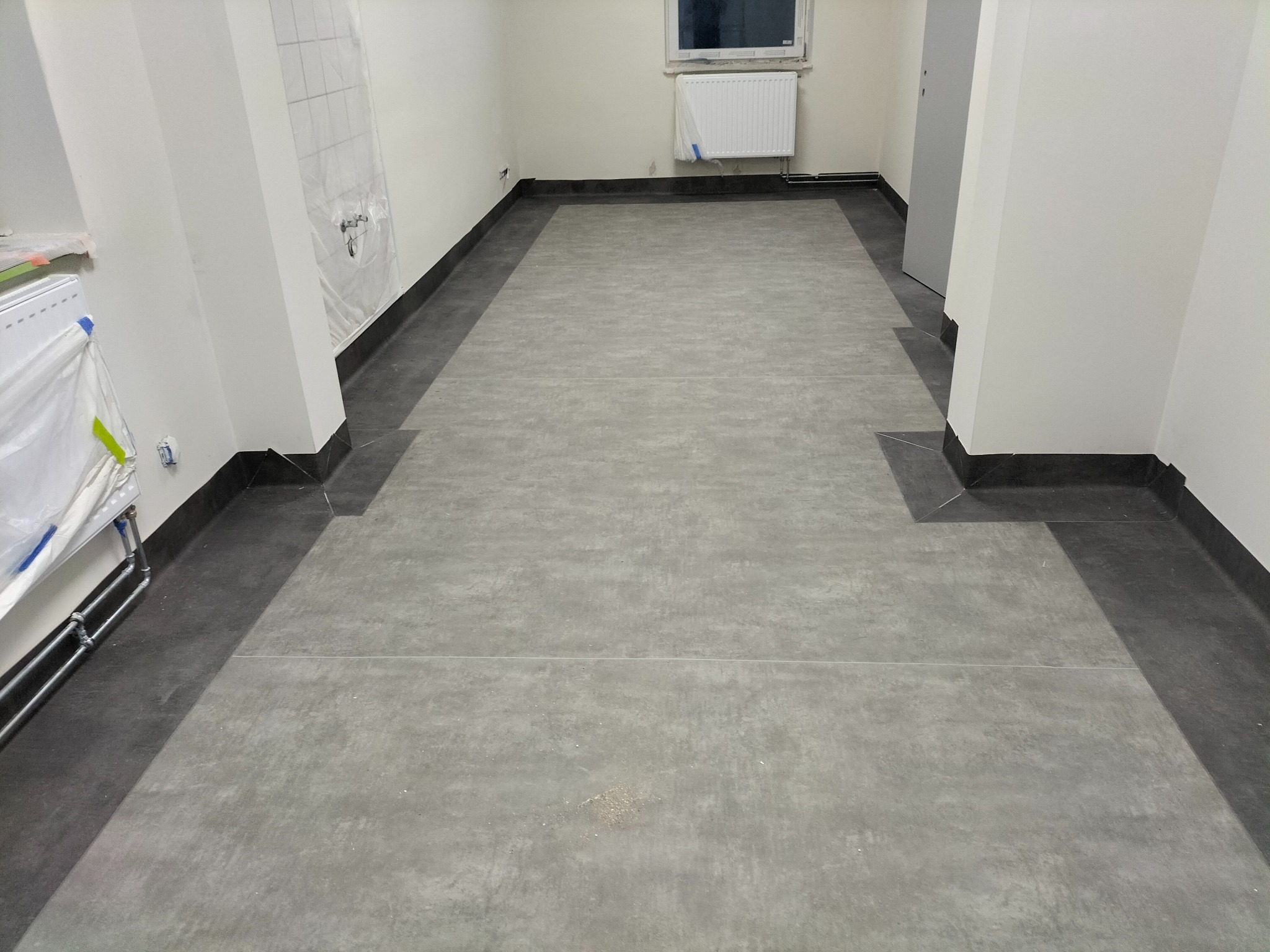 Szara podłoga z ciemnymi listwami przypodłogowymi w nowo wykończonym pomieszczeniu, widoczne pionowe rury i grzejnik zabezpieczone folią malarską.