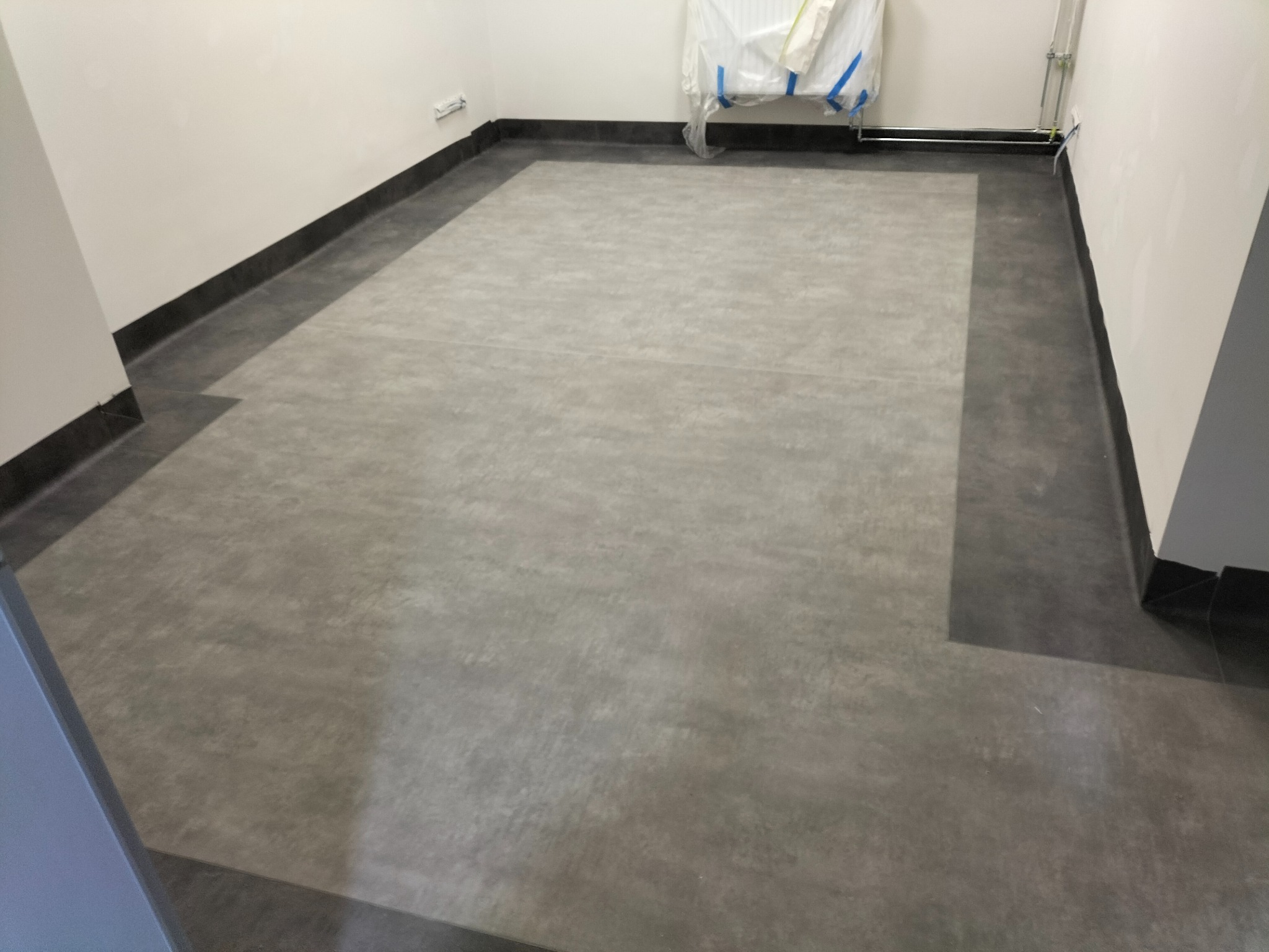 Wnętrze z nową, szarą wykładziną podłogową z ciemną listwą przypodłogową, zabezpieczone meble w tle.