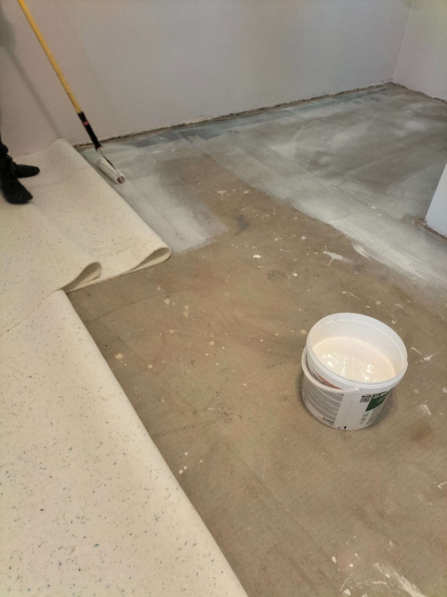 Przygotowanie podłoża pod wykładzinę: gruntowanie wałkiem. Rolka wykładziny obok, wiadro z gruntem. Widoczne ślady farby na podłodze.