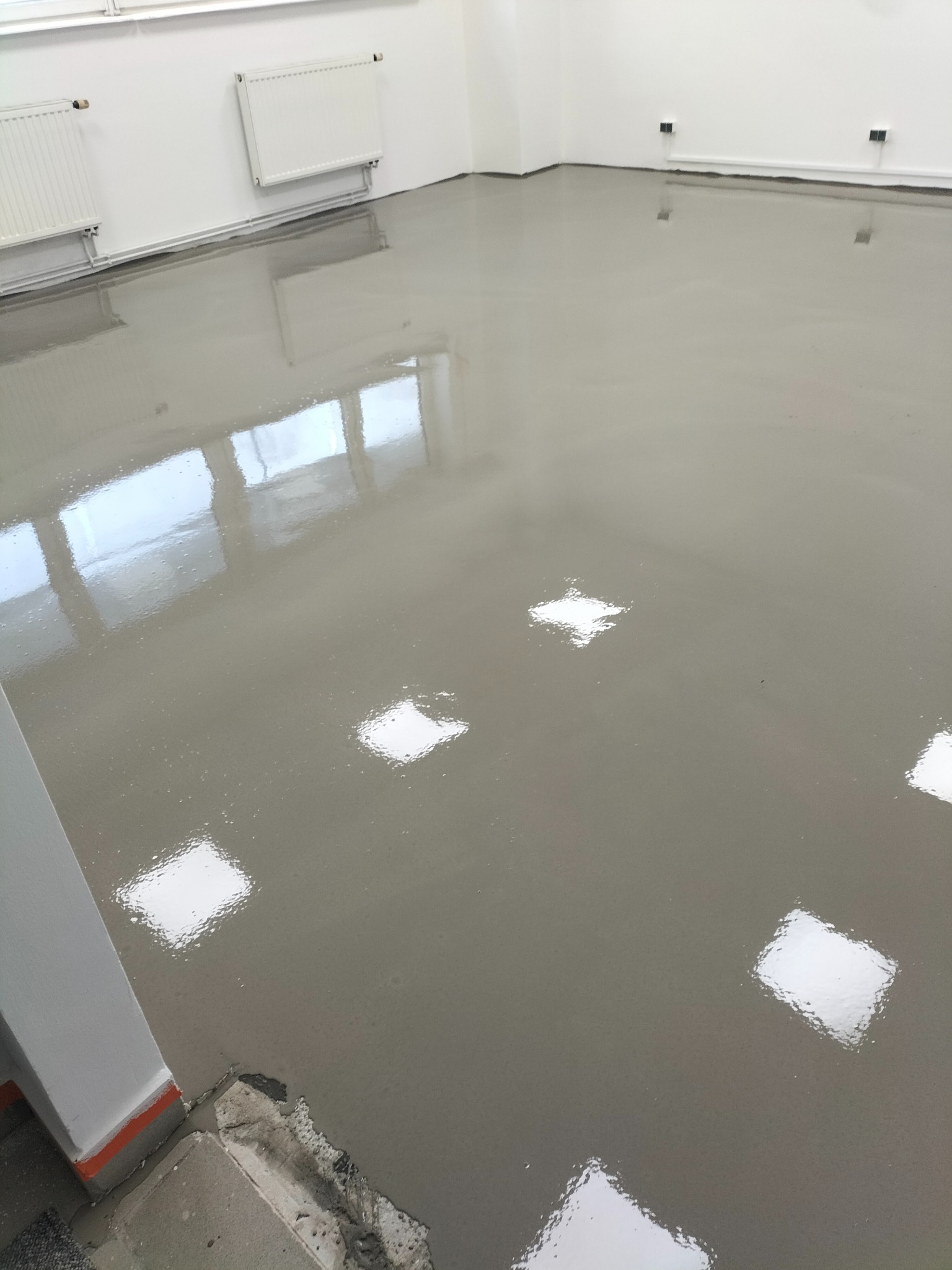 Błyszcząca, szara posadzka żywiczna w jasnym pomieszczeniu z grzejnikami i widocznymi odbiciami światła. Fragment narożnika z uszkodzonym podłożem w dolnym rogu.