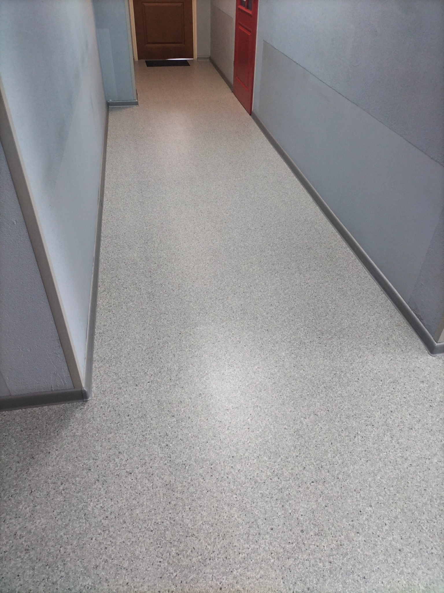 Jasnoszara wykładzina w korytarzu z czerwonymi i brązowymi drzwiami w tle. Widok z góry, prezentujący jednolitą powierzchnię i estetyczne wykończenie.