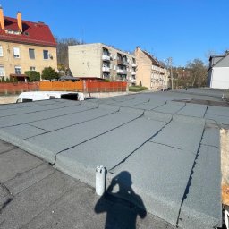 Artur Jasienowicz - Nowe, szare pokrycie dachowe z papy na płaskim dachu budynku, z widocznym cieniem osoby robiącej zdjęcie i elementami otoczenia miejskiego w tle.
