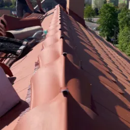 Dekarz w trakcie układania czerwonej dachówki ceramicznej na dachu domu jednorodzinnego. Widoczny komin i fragment panoramy miasta w tle. Słoneczny dzień.