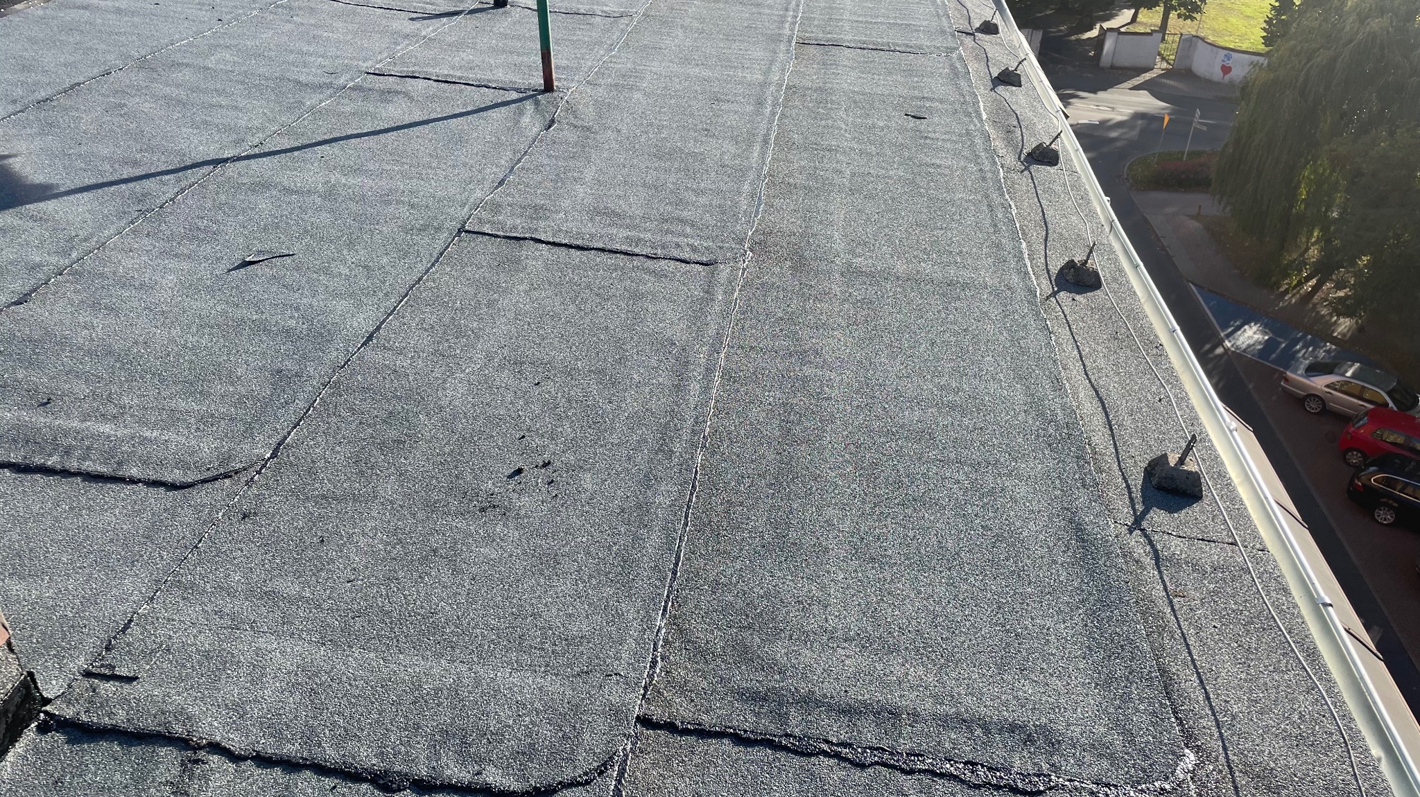 Bitumiczny dach płaski z widocznymi łączeniami papy i fragmentem rynny z uchwytami, w tle budynek i drzewa.