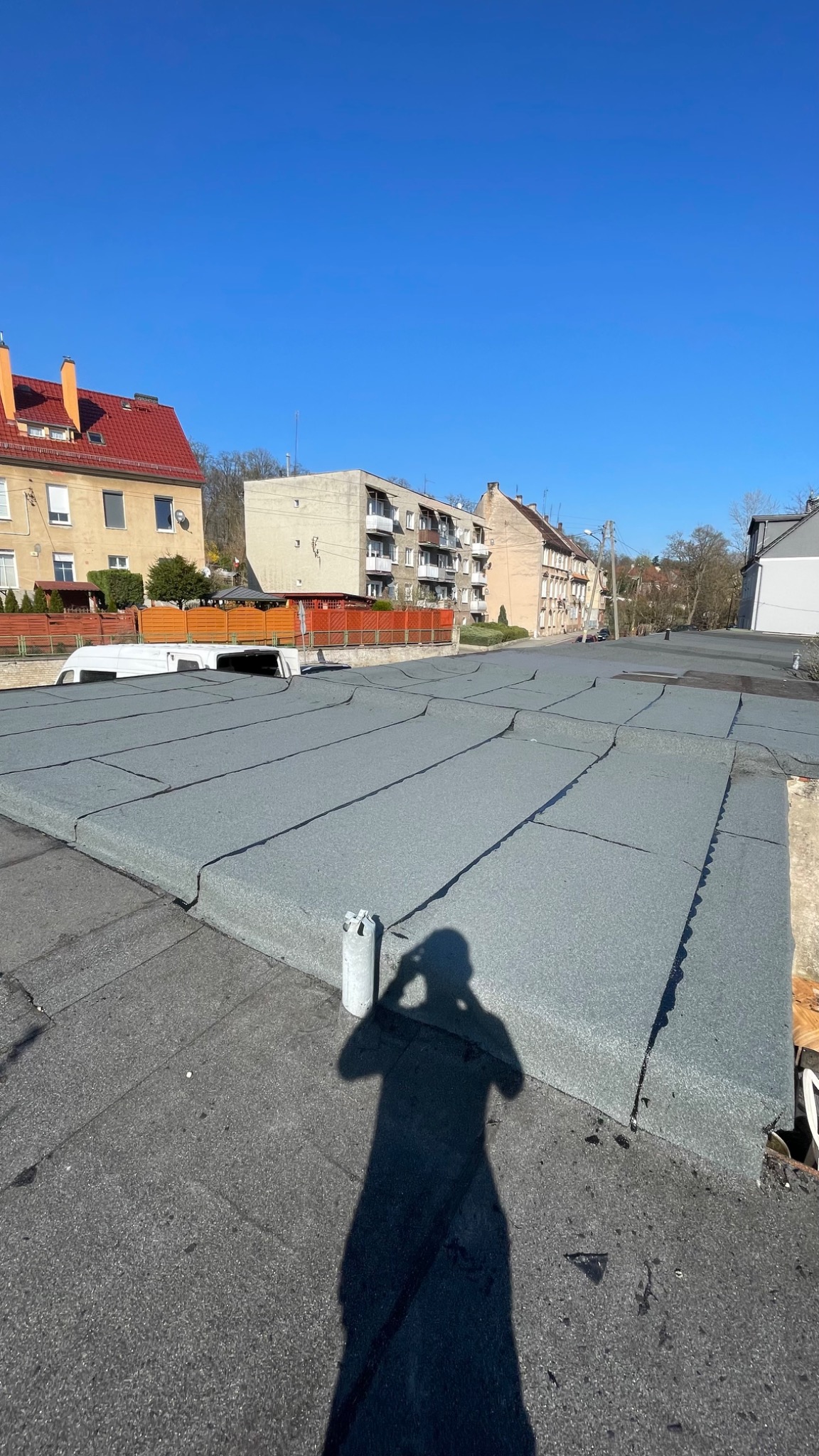 Nowe, szare pokrycie dachowe z papy na płaskim dachu budynku, z widocznym cieniem osoby robiącej zdjęcie i elementami otoczenia miejskiego w tle.