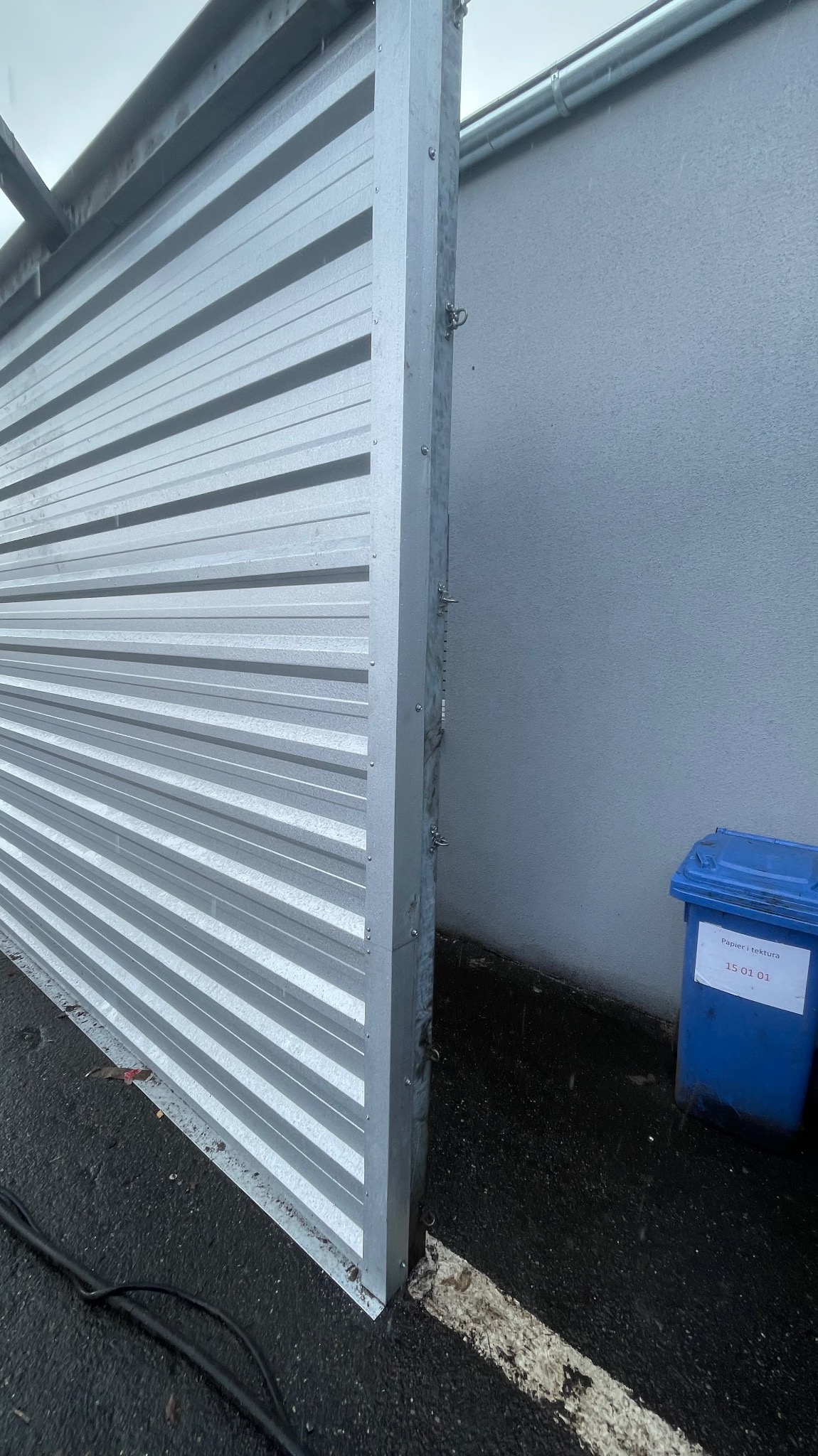 Fragment nowej, metalowej konstrukcji dachu z widocznymi poziomymi profilami, mocowanej do ściany, z niebieskim pojemnikiem na odpady w tle.