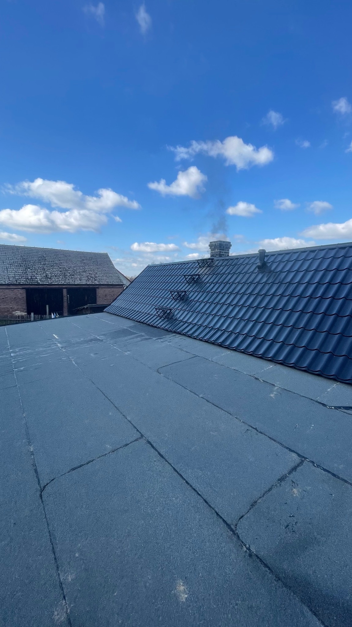Widok z góry na płaski dach pokryty papą oraz fragment dachu skośnego z blachy w kolorze grafitowym, na tle błękitnego nieba z chmurami i budynku gospodarczego.