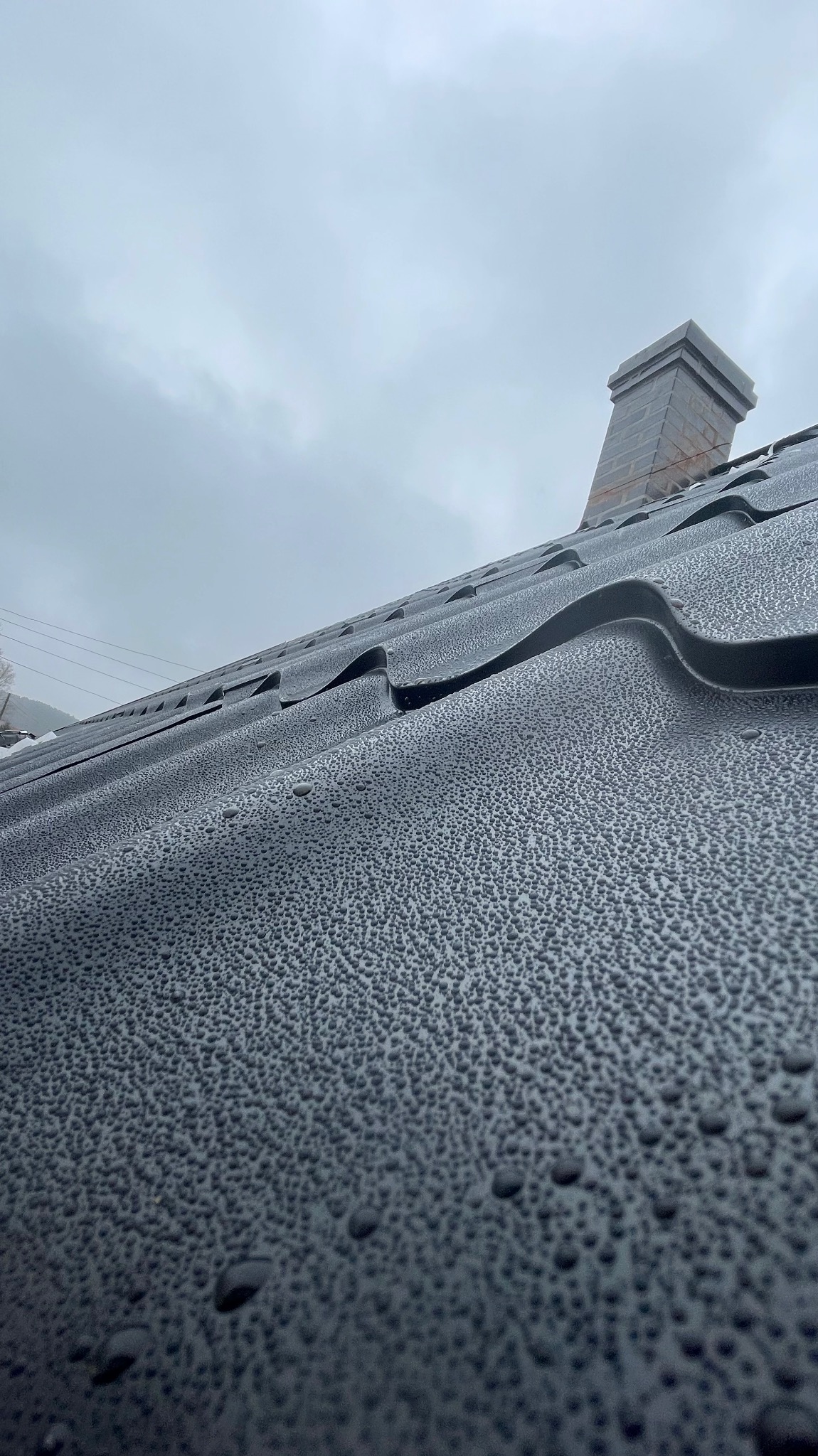 Grafitowy dach z blachy trapezowej, pokryty kroplami wody, z widocznym kominem w tle i pochmurnym niebem.