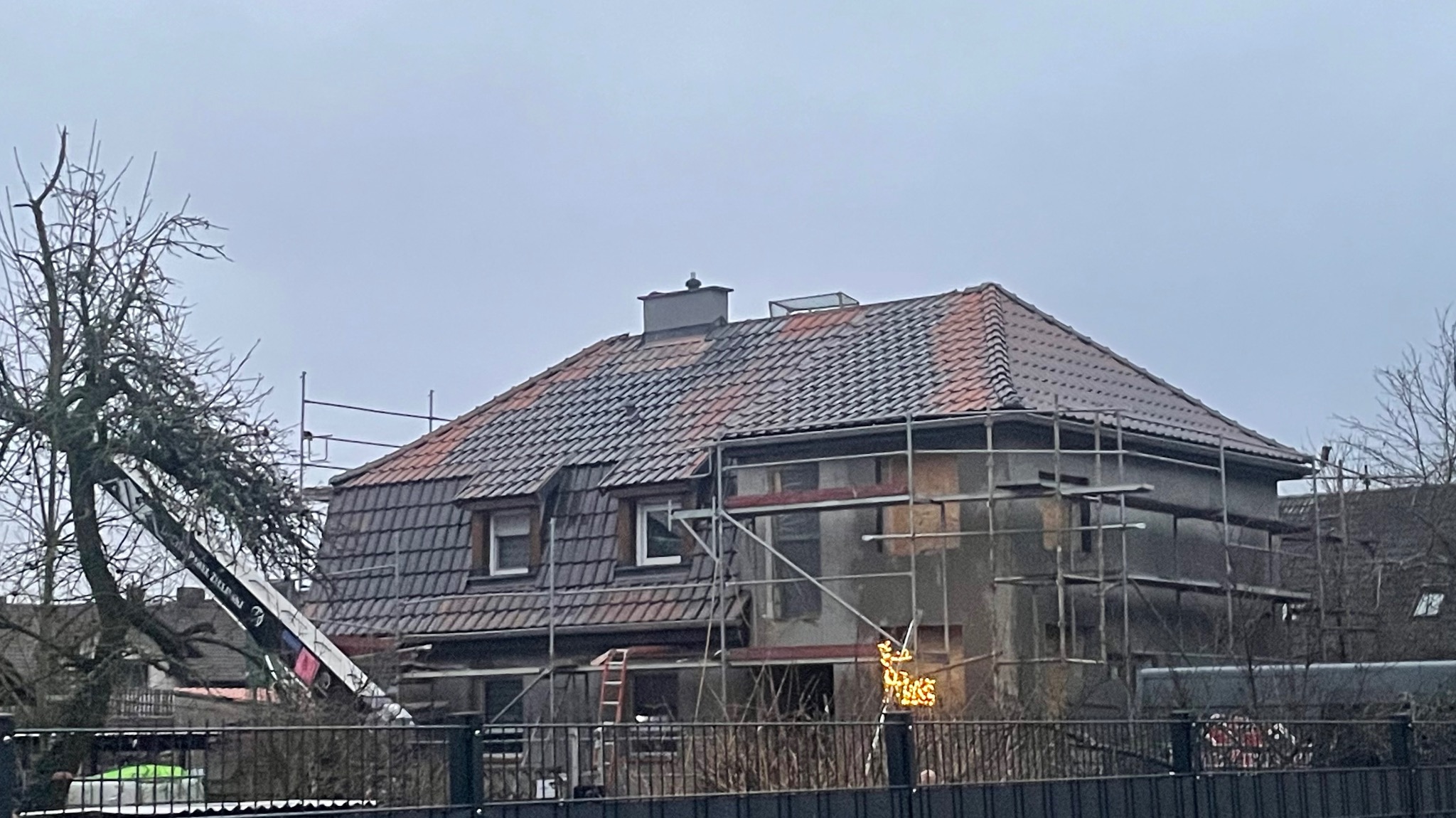 Dom w trakcie renowacji dachu z rusztowaniem i dźwigiem. Część dachu pokryta ciemną dachówką, część czerwoną. Widoczny komin i okna dachowe.