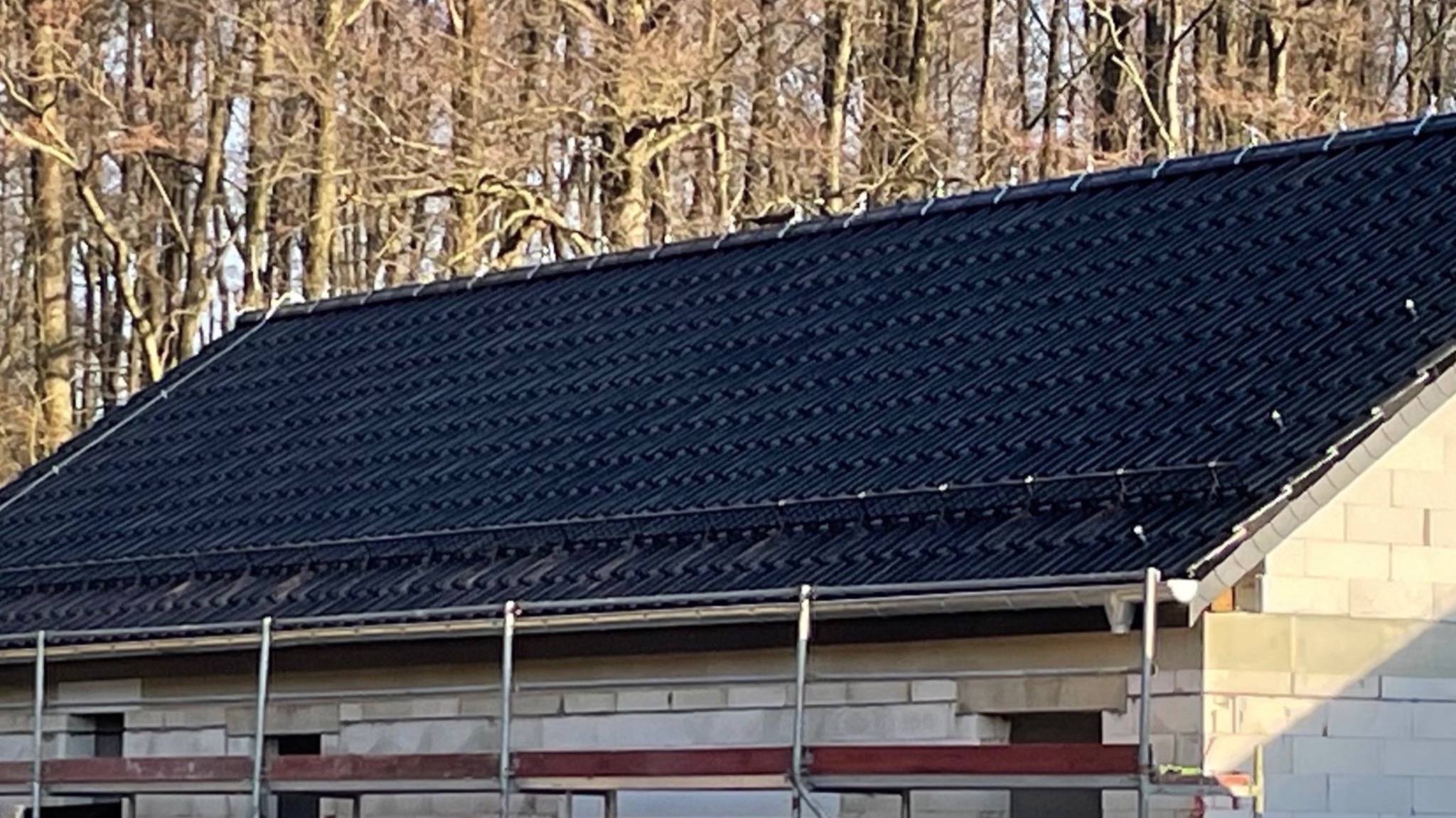 Ciemna dachówka ceramiczna na nowym budynku w trakcie budowy, widoczne rusztowania i drzewa w tle. Dach pokryty w jodełkę.