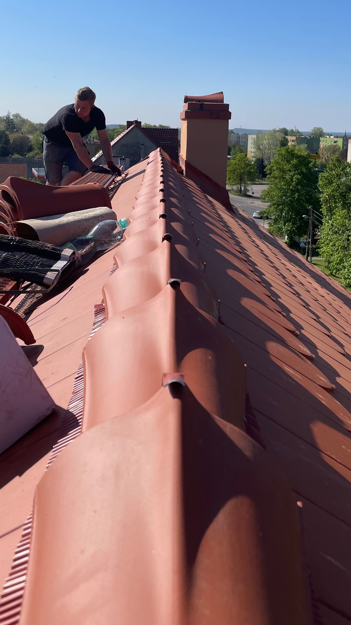 Dekarz w trakcie układania czerwonej dachówki ceramicznej na dachu domu jednorodzinnego. Widoczny komin i fragment panoramy miasta w tle. Słoneczny dzień.