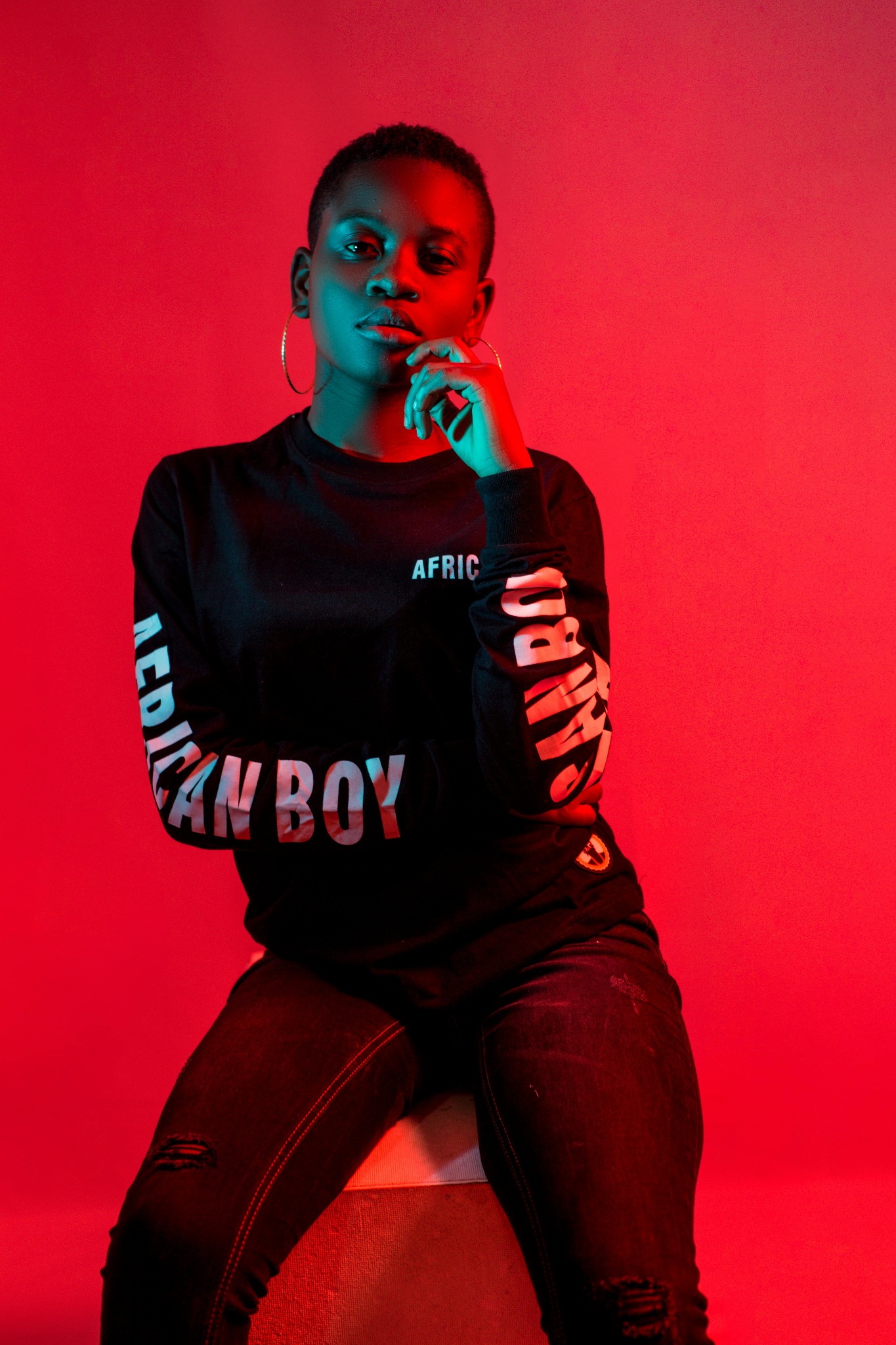 Portret młodej kobiety o krótkich włosach w czarnej bluzie z napisem 'AFRICAN BOY' na rękawach, pozującej na czerwonym tle z niebieskim oświetleniem na twarzy i ręce.