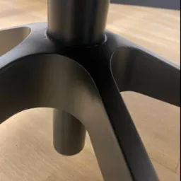 Baza czarnego, metalowego krzesła biurowego na drewnianej podłodze. Widok z dołu, skupiony na nodze krzesła i fragmencie podłogi.