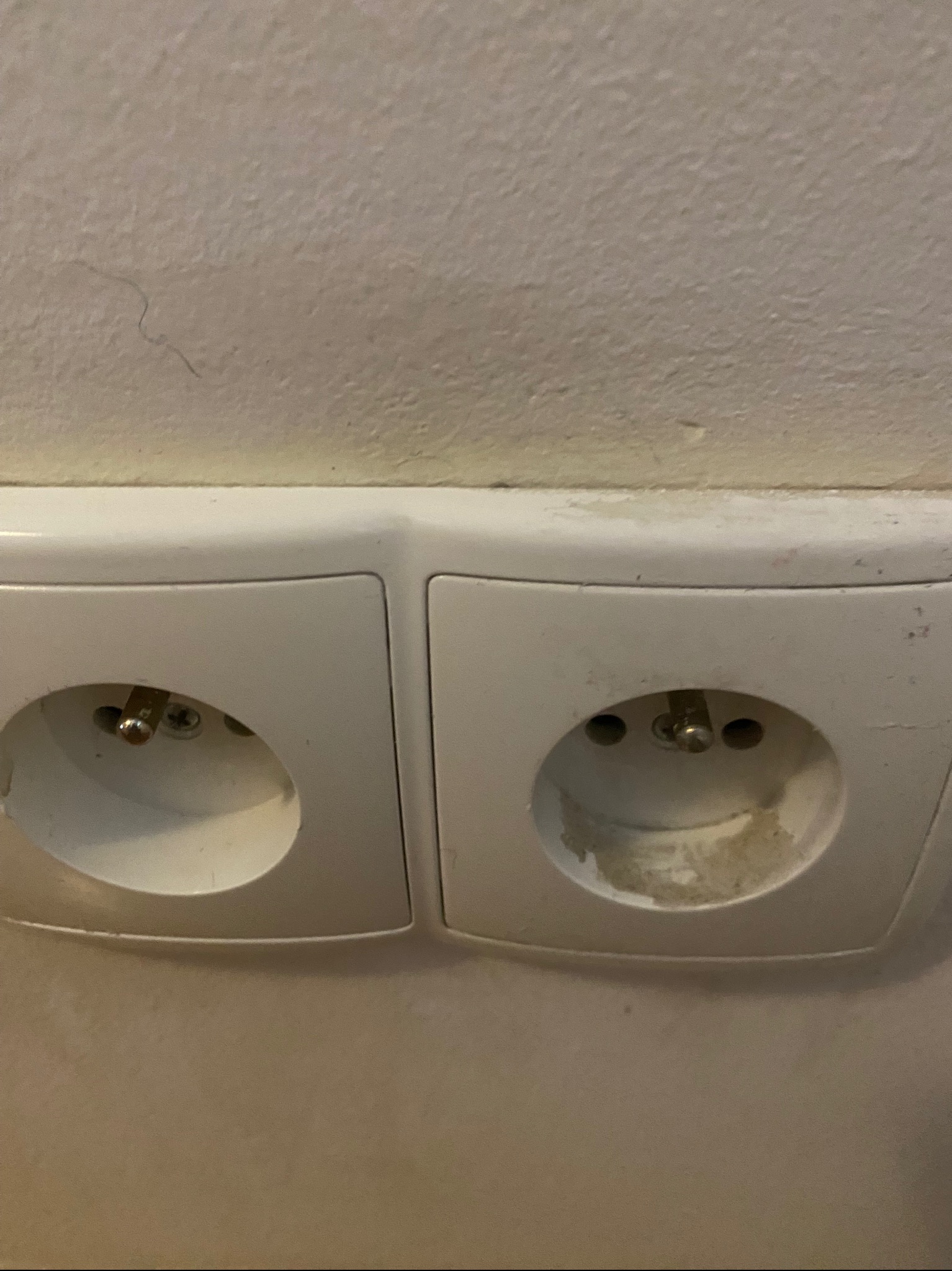Dwa białe, brudne gniazdka elektryczne na ścianie, widoczne ślady użytkowania i zabrudzenia wokół otworów.