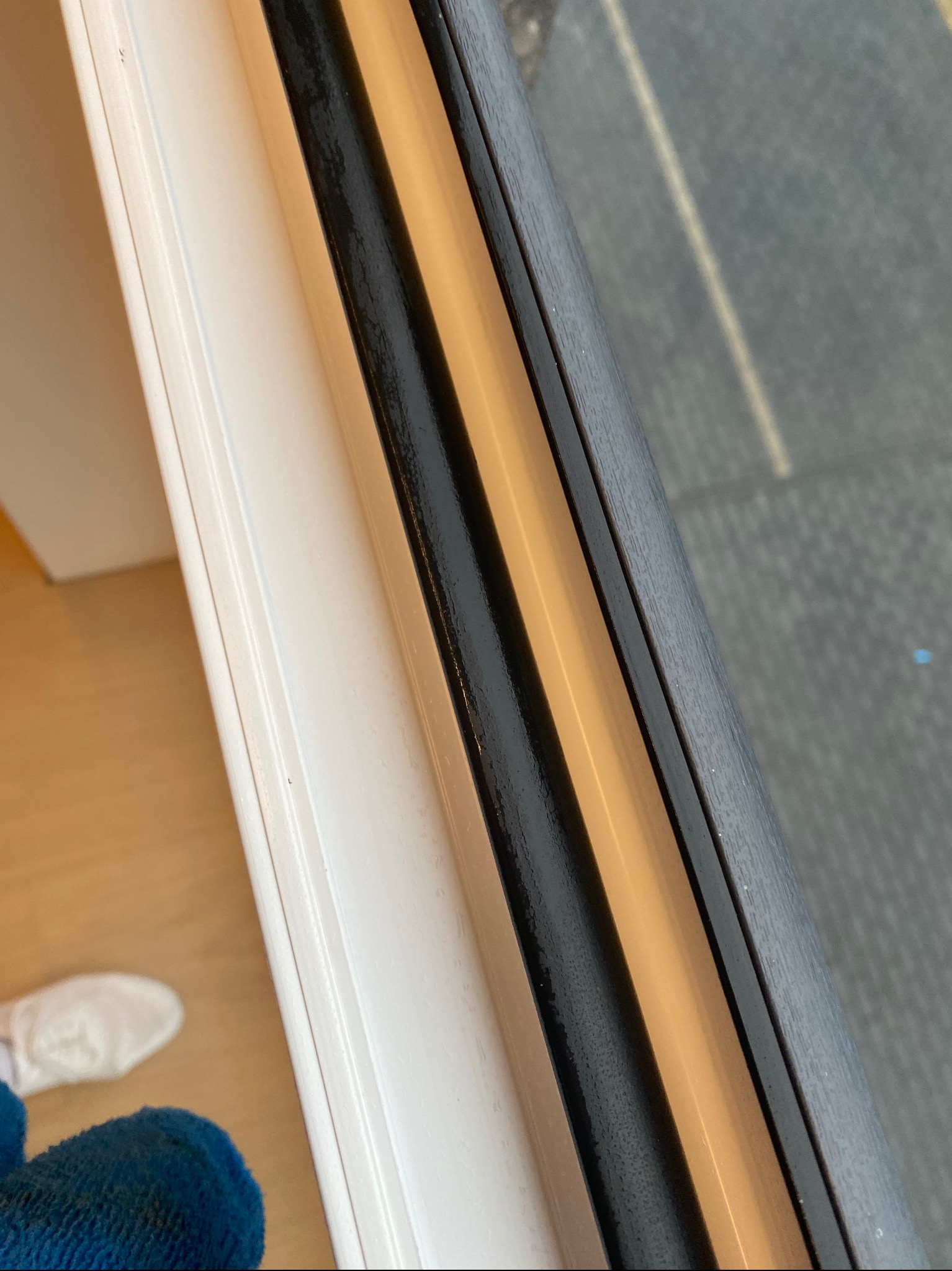 Szczegółowe ujęcie ramy okiennej z widocznymi smugami i zaciekami po myciu, ukazujące kontrast między czystą a niedoczyszczoną powierzchnią, z fragmentem podłogi i buta w tle.