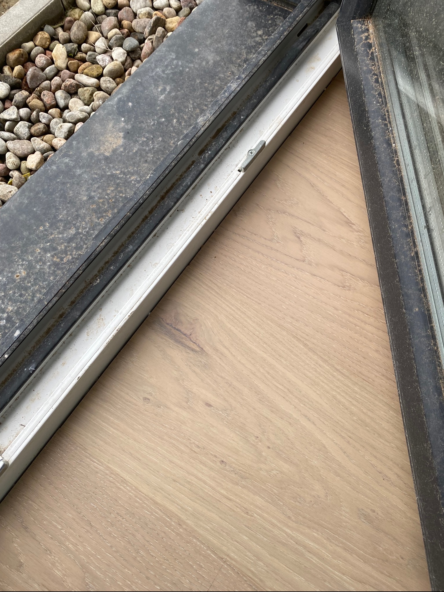 Brudna rama okienna z widocznymi śladami kurzu i zabrudzeń, przylegająca do drewnianej podłogi i kamiennego parapetu zewnętrznego.