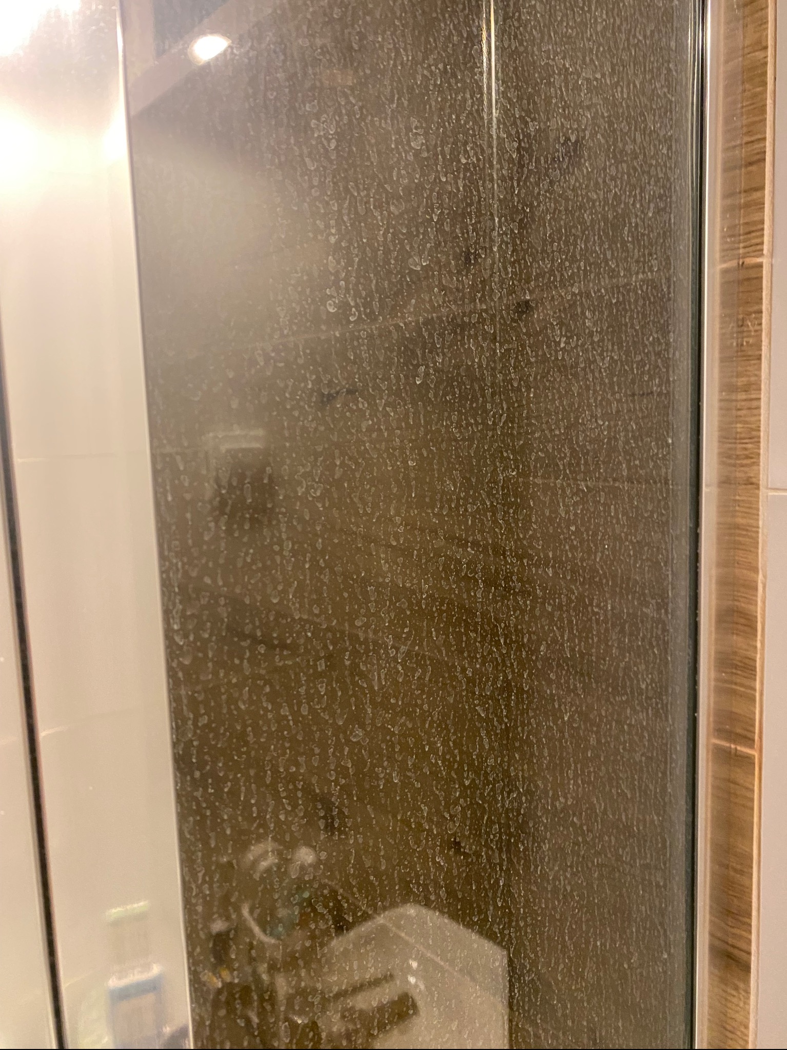 Silnie zabrudzona kabina prysznicowa z widocznymi zaciekami i osadami z kamienia na szklanych drzwiach, z fragmentem białej ściany po lewej stronie i drewnopodobnym wykończeniem po prawej.