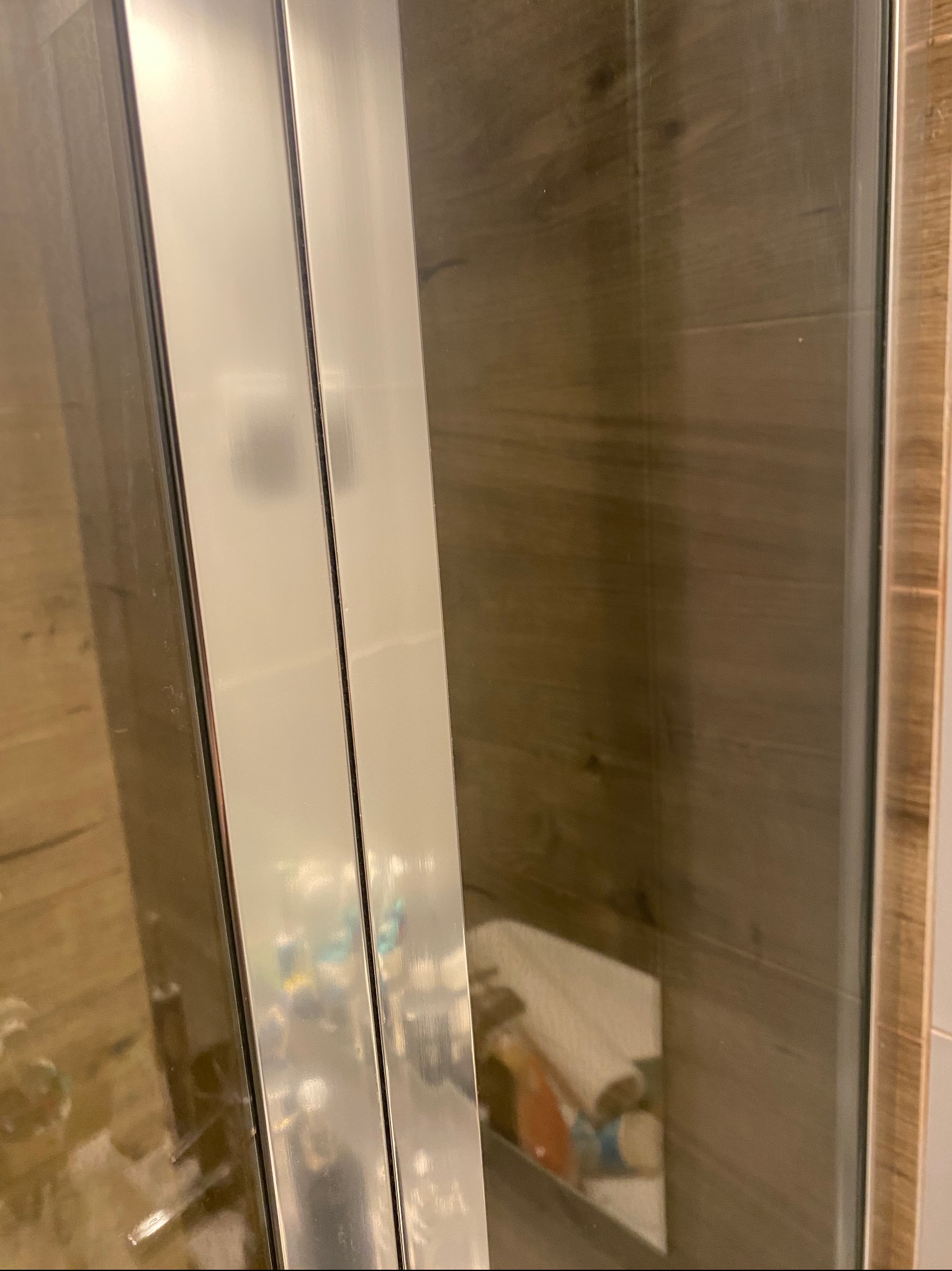 Szklana kabina prysznicowa z widocznymi śladami zacieków i zabrudzeń na powierzchni, odbijająca nieporządek w łazience, z drewnopodobną okładziną w tle.