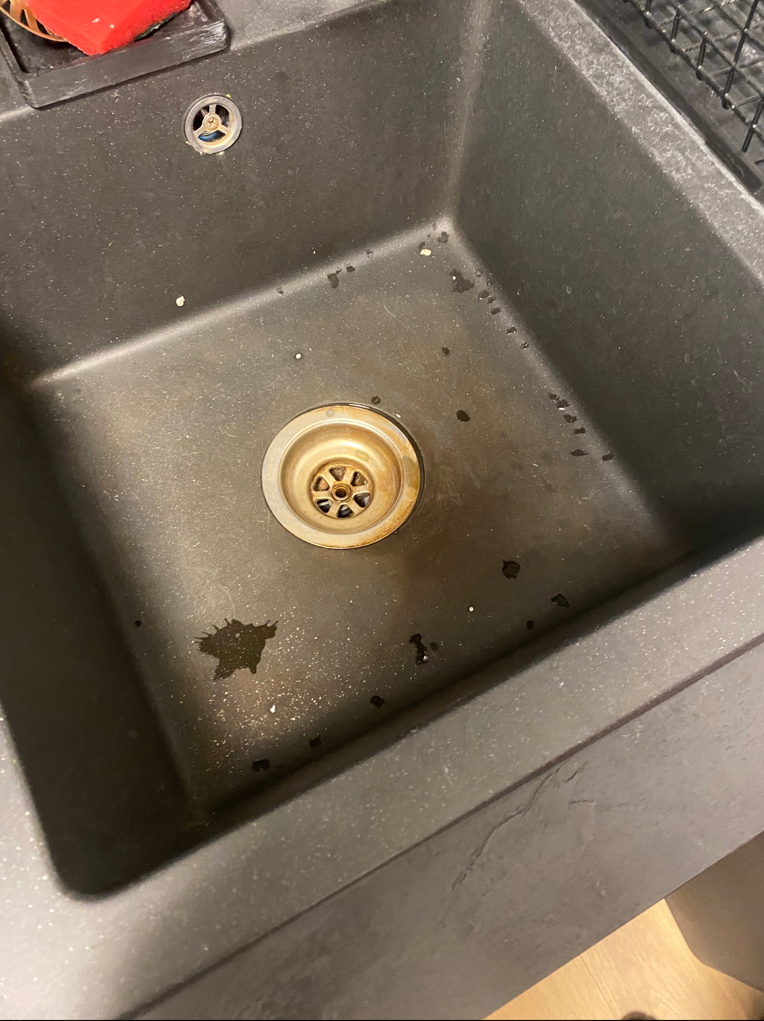 Brudny, granitowy zlew kuchenny z widocznymi plamami i resztkami jedzenia w okolicy odpływu. W rogu czerwona gąbka. Zlew wymaga czyszczenia.