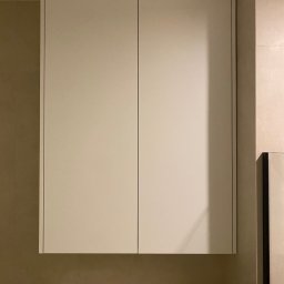 AURA JAN PIETRZAK - Zamknięta, kremowa szafka wisząca z dwoma drzwiczkami w nowoczesnej łazience z beżowymi płytkami.